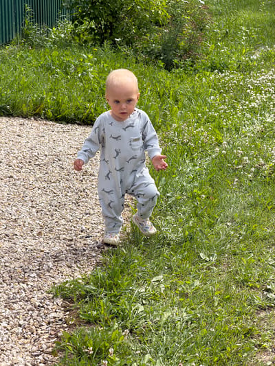 A young child with blonde hair and blue eyes, wearing a light blue long-sleeved onesie with a grey whale pattern and white sneakers, stands on a gravel path bordering a grassy area in Борисьево Днп, Russia. The child is facing forward, with their right hand slightly raised. The background consists of lush green grass, some with small white flowers, and a dark green fence with vertical slats. The lighting suggests it is daytime, with sunlight casting shadows.