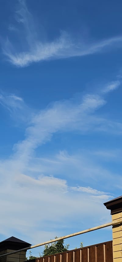 The image depicts a bright, clear sky with scattered wispy white clouds. The sky is a vivid blue, indicating a sunny day. The perspective is from below, looking upwards, suggesting the photo was taken outdoors. On the right side of the frame, part of a building's exterior is visible. This includes a dark brown, ribbed overhang or awning, and above that, a beige brick wall. A rusty metal pole runs diagonally across the frame, positioned in front of the sky and building elements. Green foliage from a tree is also visible peeking out from behind the building structure. The overall impression is of a serene daytime scene, possibly in a semi-urban or suburban setting. There is no visible text.
