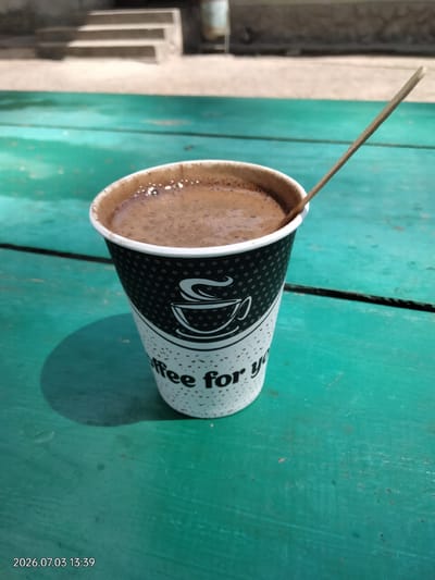 A close-up shot features a disposable paper cup filled with a dark beverage, likely coffee, resting on a weathered, teal-colored wooden surface. A slender wooden stirrer is inserted into the drink, angled towards the upper right of the frame. The cup is adorned with a repeating pattern of small white squares and a stylized white coffee cup logo, along with the partial text "ffee for y". A distinct shadow is cast by the cup onto the wooden table. In the blurred background, concrete steps ascend towards a building. The lower left corner of the image displays a timestamp: "2026.07.03 13:39". The scene is captured outdoors, suggesting a casual outdoor setting. The location is identified as Novyi Myliatyn, Ukraine.