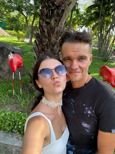 This is a selfie taken in a park-like setting in Thuận Nam, Vietnam. The image features a couple posing for the camera. The woman is in the foreground, wearing a white top and a decorative choker, with sunglasses on and her lips pursed. She has dark hair pulled back. Next to her, the man is looking directly at the camera. He is wearing a black t-shirt with a galaxy print. In the background, there are trees, green grass, and a rock formation. Two red flamingo lawn ornaments are visible, one to the left and another partially visible to the right of the couple. The lighting suggests it is daytime.