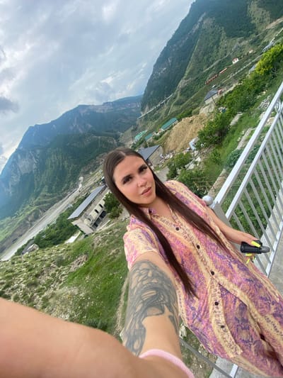 A woman with long, dark hair and a patterned dress takes a selfie on a balcony overlooking a mountainous landscape. The woman is holding a black object with a neon green tip in her right hand. The mountainous terrain features green vegetation, scattered buildings, and a landslide. The sky is cloudy. The location appears to be Chalda, Russia, based on the prompt.