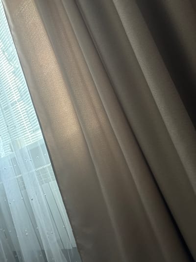 The image displays a close-up, vertical view of draped curtains. On the left side, a sheer white curtain with small, scattered white beads is visible, layered in front of a window with horizontal blinds. The main subject of the image, occupying the right two-thirds, is a heavier, textured curtain in a muted taupe color. This curtain is heavily pleated, creating deep shadows and highlights that emphasize its fabric texture and folds. The lighting suggests it is daytime, with light filtering through the window and illuminating the lighter curtain and the front folds of the taupe curtain. There is no indication of people, actions, or any context related to the city of Kinel'-Cherkasy, Russia. The scene is static and focuses solely on the textile surfaces and the interplay of light and shadow.