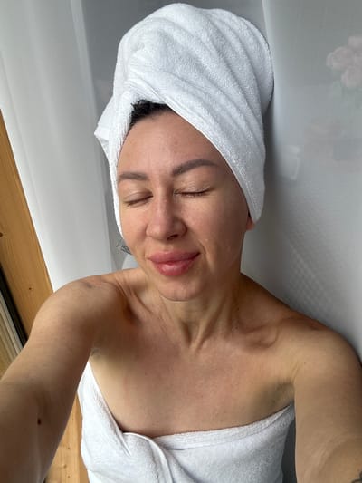 A woman is lying on her side, her eyes closed and her head tilted back. She is wearing a white towel wrapped around her head like a turban and another towel draped over her chest and shoulder. Her skin is slightly damp, suggesting she may have just showered or is undergoing a spa treatment. A label is visible on the towel on her chest, which reads "HOF COSMETICS". The background appears to be a bathroom or spa-like setting, with a white surface above and wooden paneling below. The overall mood is one of relaxation and self-care. The lighting suggests it is daytime.