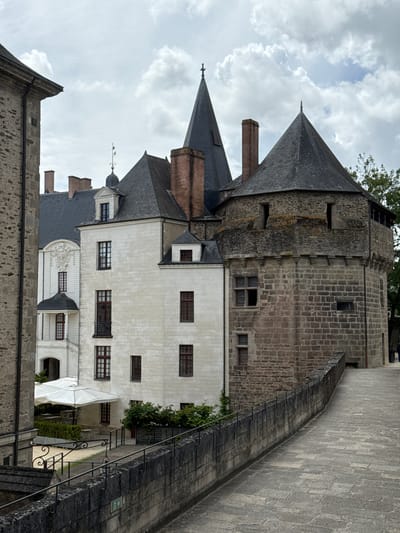 The image captures a view of the Château des Ducs de Bretagne in Nantes, France, on a cloudy day. In the foreground, a cobblestone pathway with a stone wall and railing extends to the right. The wall is topped with a metal railing. Further into the background, the Château's architecture is visible, featuring a large stone tower with a conical roof on the right, and a white building with a mansard roof and a pointed spire to its left. Several windows with red trim are visible on the white building, and a small balcony protrudes from its facade. Behind the white building, a darker, older section of the castle can be seen with a steep, dark roof. Green foliage is present near the base of the white building and further back in the background. A white canopy is set up over some outdoor seating areas. The sky is filled with scattered clouds.