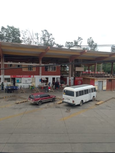 The image captures a scene in Municipio Tarija, Bolivia, displaying a red-brick building with a covered area, likely a commercial or transportation hub. The building houses businesses like "PATIO DE COMIDAS" (Food Court) and "CASCADA." People are gathered outside the building, possibly customers or travelers. A red station wagon and a white minibus are parked in a paved area marked with yellow parking barriers. The background shows trees and an overcast sky. Utility lines stretch across the top portion of the image.