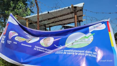 This media file captures a blue public health awareness banner displayed outdoors in Tchere, Democratic Republic of the Congo. The banner is slightly curved, indicating it's made of flexible material, and is affixed to a wire fence, with barbed wire visible across the top and in the background. Beyond the fence, there are wooden structures, corrugated metal roofs, and green foliage, all under a bright blue sky, suggesting daytime.

The banner presents health information in French, utilizing pictograms to illustrate symptoms and recommended actions. From left to right, the images depict: a person lying down, corresponding to "Apparition brutale de la fièvre" (Sudden onset of fever); a person with "Diarrhée" (Diarrhea); a person experiencing "Vomissements" (Vomiting); and a person with "Saignement inhabituel par la bouche et le nez" (Unusual bleeding from the mouth and nose).

Following these symptom illustrations, a large arrow points to a circular graphic showing a building labeled "CENTRE DE SANTE" (Health Center), topped with a prominent green checkmark. The accompanying instruction reads: "Allons immédiatement dans un centre de santé le plus proche." (Let's go immediately to the nearest health center.)

Partial text visible at the top left indicates "MINISTERE DE LA SANTE PUBLIQUE" (Ministry of Public Health) and "Programme National de Santé Publique" (National Public Health Program). A logo for the Ministry of Public Health is also present. The bottom right corner credits "Joseph Kisuki." The banner functions as a crucial tool for public health communication in the region.