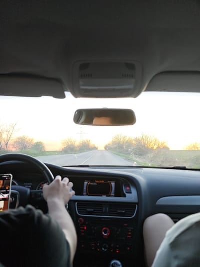 The view from inside a car shows a road stretching into the distance, likely taken in the vicinity of Gorni Tsibar, Bulgaria, given the rural landscape and the possible presence of electrical pylons. The driver's hands are on the steering wheel, and a mobile phone is visible on the dashboard. The dashboard of the car displays an informational screen. The passenger's leg is partially visible in the bottom right of the frame. The lighting indicates either sunrise or sunset.