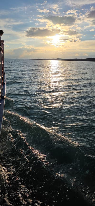 This is a vertical, medium shot photograph taken from the side of a vessel traveling across a body of water. The sun is setting, casting a golden sheen across the rippling surface of the water, which is dark blue in color. The sun is low in the sky, partially obscured by clouds, and is reflected on the water. The horizon line is visible in the distance, with a dark, undulating landmass on the far side of the water. The left side of the photograph shows the edge of the vessel, with a railing and some rope visible. The overall impression is one of tranquility and natural beauty. The location is the Issyk-Kul Lake, near the city of Pristan'-Przheval'sk, Kyrgyzstan.