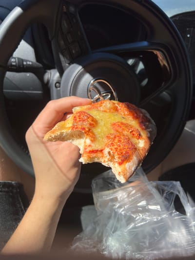 A hand is holding a slice of cheese and tomato pizza in front of a car's steering wheel. The steering wheel has buttons on the right side and a circular emblem in the center. The background is dark, suggesting the interior of a car. A crumpled plastic bag is visible in the lower right corner. The lighting indicates it is daytime, with sunlight casting shadows. The pizza appears to have a slightly burnt crust. The hand holding the pizza has visible arm hair. The scene is captured from a first-person perspective, as if the viewer is the one holding the pizza. The location is not specified in the image.