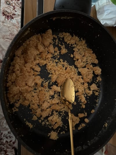 A close-up, overhead shot shows a black frying pan filled with a crumbly, light brown mixture and a golden spoon resting in the mixture. The pan is on a wooden surface, and to the left of the pan, a patterned fabric is visible. To the upper right, a white bag with a clear lid is also visible. The image appears to be a still life, possibly depicting a culinary preparation. There is no information available about the specific location of Oberjettingen, Germany.