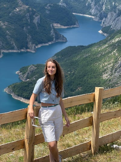 A young woman stands near a wooden fence, holding a pair of sunglasses in her right hand. She is wearing a light blue cropped shirt and off-white shorts, with a green belt. Her long brown hair is partially blowing in the wind. Behind her, a vast, blue lake is nestled within a canyon, surrounded by steep, green, forested mountains. The scene appears to be a scenic overlook. The location is identified as Dubljevići, Montenegro, specifically overlooking the Piva Lake.