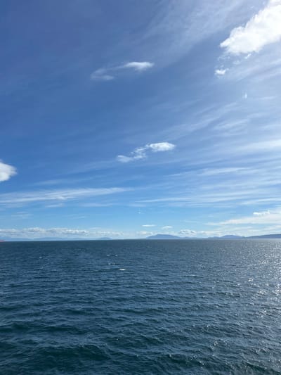 The image displays an expansive marine vista under a partly cloudy sky within the Southern Gulf Islands Electoral Area, Canada. The upper half of the frame is dominated by a clear blue sky with scattered white clouds, including wispy cirrus and smaller cumulus formations. The lower half features dark blue water exhibiting surface ripples and reflecting sunlight, particularly on the right side. Along the distant horizon, several landmasses, appearing as vegetated islands or coastal outlines, are visible. A large, reddish-orange vessel, likely a ship or ferry, is partially discernible on the far left horizon. No individuals or specific actions are observed.