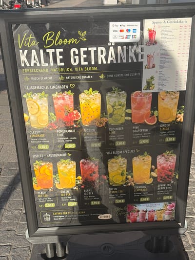 This is a close-up, eye-level, outdoor shot of a black menu board for "Vita Bloom" drinks. The board is positioned on a paved stone sidewalk, suggesting an urban environment like Würzburg, Germany, possibly near a cafe or shop entrance. The menu is predominantly black with white and yellow text, featuring images of various drinks.

The top section displays "Vita Bloom KALTE GETRAENKE" (Vita Bloom Cold Drinks) with the tagline "ERFRISCHEND. NATÜRLICH. VITA BLOOM. OHNE KÜNSTLICHE ZUSÄTZE" (Refreshing. Natural. Vita Bloom. Without Artificial Additives.). Below this are two smaller icons and text reading "FRISCH GEMACHT" (Freshly Made) and "NATÜRLICHE ZUTATEN" (Natural Ingredients).

The main body of the menu is divided into sections: "HAUSGEMACHTE LIMONADEN" (Homemade Lemonades) and "EISTEES - HAUSGEMACHT" (Homemade Iced Teas).

The "Homemade Lemonades" section features images of six drinks:
- CLASSIC LEMONADE (0.5L 3,50 €)
- POMEGRANATE LIME (0.5L 3,90 €)
- PASSION FRUIT LEMONADE (0.5L 3,90 €)
- CUCUMBER LIME (0.5L 3,50 €)
- PINK GRAPEFRUIT (0.5L 3,90 €)
- GINGER LEMON (0.5L 3,50 €)

The "Homemade Iced Teas" section displays four drinks:
- PEACH ICE TEA (0.5L 3,50 €)
- GREEN ICE TEA (0.5L 3,50 €)
- BERRY ICE TEA (0.5L 3,90 €)
- SPARKLING BERRY LIME (0.5L 3,90 €)

At the bottom of the "Homemade Iced Teas" section, there's a box for "EXTRAS FÜR DEIN GETRÄNK" (Extras for your Drink) with options like "Extra Ingwer", "Extra Beeren", "Extra Zitrone", "Extra Minze", and "Extra Honig", with a price of "je +0,50 €" (each +0.50 €).

To the right of the menu board, there is a smaller, separate menu titled "Smoothies & more! Speise- & Getränkekarte" (Food & Drinks Menu). This section lists "Smoothies," "Milkshakes," and "Säfte" (Juices) with various flavors and prices in Euros for 300ml, 400ml, and 500ml sizes. It also includes payment logos such as G Pay, Samsung Pay, Visa, Mastercard, American Express, Diners Club, JCB, and Maestro.

In the background, partially visible at the top of the image, is a blurred image of a bicycle. The lighting suggests it is daytime, with sunlight creating some glare on the menu board. The overall condition of the menu board appears good, with no visible damage.