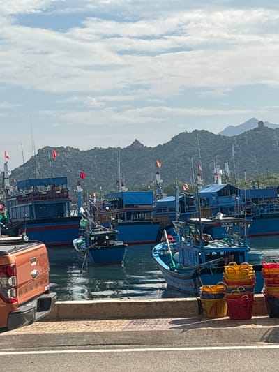The image captures a vibrant daytime scene at a harbor in Ninh Chu Ward, Vietnam, under a sky filled with soft, white clouds. The setting features a row of numerous fishing boats, primarily painted in shades of blue with some red accents, moored along a concrete quay. Many of these boats display small red flags fluttering from their masts.

On the quay itself, stacks of colorful baskets, predominantly red and yellow, are neatly arranged, suggesting their use for fishing or transporting goods. In the immediate foreground, a partial view of an orange pickup truck is visible, with the brand name "RANGER" clearly legible on its tailgate. The water in the harbor is calm, reflecting the boats and the overhead clouds.

In the background, a range of lush, green mountains rises, providing a striking natural backdrop to the bustling port. The scene depicts a typical working harbor, likely a fishing village or town, with boats at rest or preparing for activity. No specific individuals are clearly visible, but the presence of the boats and equipment implies ongoing human activity related to the fishing industry. The bright daylight suggests it is late morning or early afternoon.