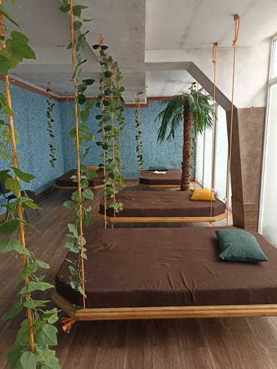 This media file depicts a tranquil relaxation area with a tropical theme. The primary subjects are several low-profile, brown-covered daybeds suspended from the ceiling by orange ropes, creating a swing-like effect. These daybeds are adorned with cushions, one a vibrant yellow and another a deep teal, adding pops of color to the otherwise muted palette.
The setting is indoors, suggested by the concrete ceiling and the presence of artificial greenery. Lush, green artificial vines hang down from the ceiling, entwined with the suspension ropes, enhancing the tropical ambiance. In the background, a tall artificial palm tree stands prominently, further contributing to the exotic feel. The walls are covered in a light blue mosaic tile, and the floor is made of polished wooden planks, adding to the resort-like atmosphere.
There are no people visible in the image, indicating a quiet, unoccupied space. The activity taking place is implied: relaxation, lounging, or possibly spa treatments. The lighting appears to be natural, possibly from large windows or skylights, though they are not clearly visible, suggesting it could be daytime. The overall emotion evoked is one of peace, serenity, and escape.
The location cues, such as the artificial palm tree and tropical plants, along with the blue tiled walls, suggest a spa, resort, or a dedicated relaxation room. There is no visible text in the image. The specific location of Сонячне, Ukraine, provides geographical context for this type of recreational or wellness facility.