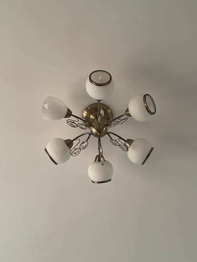 The image shows a close-up, overhead view of a brass chandelier with six white glass shades attached to it. The chandelier is mounted on a white ceiling. The shades are of two different shapes: three are round globes, and three are more conical. Some of the shades have a metallic trim around the opening. Between the shades, there are decorative elements that appear to be clear crystals. The chandelier is illuminated, as evidenced by the light visible within some of the shades. The background is a plain white ceiling, showing some subtle texture. The location is identified as Novogorelovo, Russia.