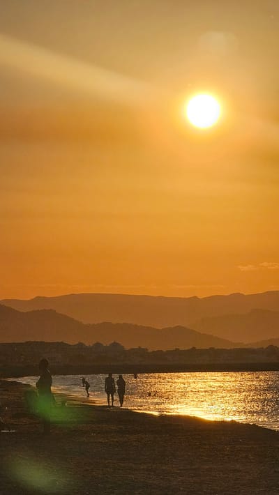 The image captures a serene sunset scene at Deveses beach in Spain. The sun, a bright orb, hangs high in the sky, casting a warm, golden glow across the landscape. The sky is a gradient of vibrant orange, fading into softer hues as it reaches the horizon. Silhouetted against this luminous backdrop are several people walking along the shore. Two figures stand close together near the water's edge, while another person stands further back on the beach, looking out towards the sea. In the distance, layers of mountains create a dramatic silhouette against the warm sky, and a strip of buildings suggests a coastal town. The sea reflects the sunlight, creating a shimmering pathway of golden light that stretches across the water. The foreground is dominated by the dark sand of the beach.