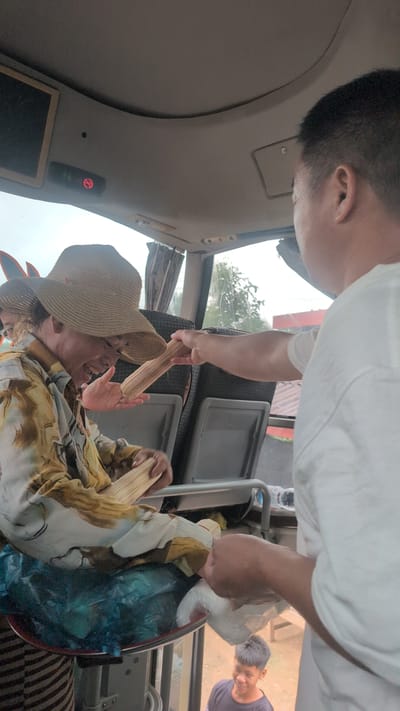 A man in a white shirt offers a bundle of wooden sticks to a person wearing a wide-brimmed straw hat inside a vehicle, likely a bus. The person in the hat, wearing a patterned shirt, is laughing and reaching out to receive the sticks. Another person, a young man in a dark t-shirt, is visible smiling in the background, outside the vehicle. The interior of the vehicle shows seats and part of the ceiling with a "no smoking" sign illuminated. The environment outside the window appears to be a rural or semi-urban area with trees and buildings.