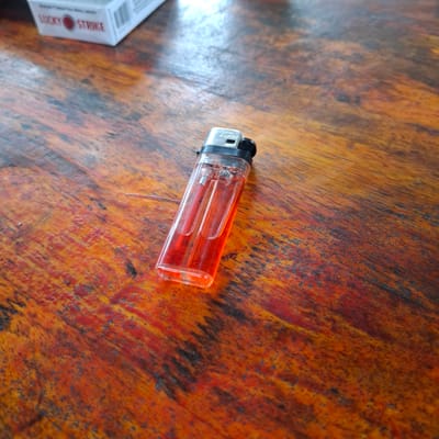 This image features a red plastic lighter placed on a wooden surface with a prominent grain pattern. A portion of a white cigarette pack with "LUCKY STRIKE" written vertically is visible on the left side of the frame, partially obscuring the wooden surface. The lighter is transparent and appears to contain fuel, with a black plastic top. The wooden surface exhibits a warm, reddish-brown hue with dark streaks and a textured finish, suggesting it might be an aged table. The lighting suggests an indoor setting, possibly with natural light illuminating the scene. There is no discernible activity or event taking place beyond the static arrangement of these objects. The overall impression is simple and direct, focusing on the objects and their immediate environment.