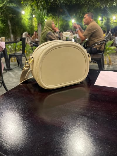 A light beige, oval-shaped handbag with a gold strap buckle rests on a dark, reflective table in what appears to be an outdoor restaurant setting in Bireuenmeunasahdayah, Indonesia. In the background, several people are seated at tables amidst lush greenery and string lights, suggesting a nighttime or evening ambiance. One person in the mid-ground, wearing a green hijab, is looking to the left, while another man in a brown shirt is looking down and holding something in his hands. The table surface shows light reflections and some minor scratches. A small pink piece of paper is visible on the table to the right of the handbag.