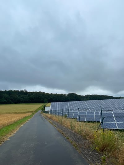 An overcast, wide shot captures a road leading to a solar panel farm located in Ottrau, Germany. The road, paved with asphalt, is wet, suggesting recent rain. To the left of the road, a field of golden wheat stretches towards a dense line of dark green trees that form the horizon. On the right, a vast expanse of solar panels is visible, arranged in neat rows and enclosed by a fence. The sky is filled with heavy, gray clouds, casting a muted light over the scene. A small, white building is situated at the edge of the solar farm, near the road. No people or animals are present in the frame.