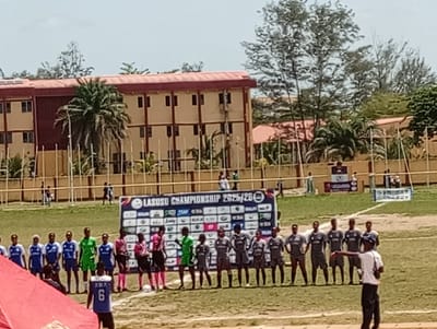 The image shows a soccer field in Isasi, Nigeria. Two teams are lined up on the field, facing each other. The team on the left is wearing blue and white jerseys, while the team on the right is wearing grey and black jerseys. There are referees in pink and black uniforms standing between the teams. A man in a white shirt and black pants is standing in front of the grey team, pointing. Behind the teams is a banner that reads "LASOSU CHAMPIONSHIP 2025/26." In the background, there is a tan building with a red roof, trees, and a fence. People are also visible in the background standing in the grass. The day appears sunny.