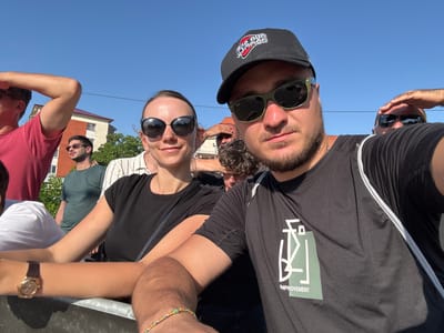 In Poti, Georgia, on a clear, sunny day, a group of people are gathered outdoors, possibly at an event or a festival. In the foreground, a young couple takes a selfie. The man, wearing a black baseball cap and sunglasses, smiles at the camera. Next to him, a woman also wearing sunglasses and a black t-shirt, poses for the photo. The couple appears to be leaning on a railing, suggesting they are observing something or waiting for an event to begin.
Behind them, other individuals are visible. A man in a red t-shirt is partially visible on the left, looking away from the camera. To the right of the couple, another man in a green t-shirt stands, his gaze directed slightly downwards. Further back, another person's arm is raised, shielding their eyes from the sun. The background shows a building with a reddish-brown roof and some trees, indicating an urban or suburban setting. The overall atmosphere seems casual and relaxed, with a hint of excitement from the gathering.