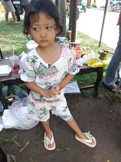 A young girl with dark curly hair stands with her legs slightly apart on a dirt surface. She wears a patterned white and green short set and pink sandals adorned with green embellishments. Behind her, a table is laden with various items, including drinks, condiments, and plastic containers. The background reveals a street scene with motorcycles and other figures.