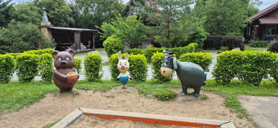 This is an outdoor, eye-level, medium shot photograph of three cartoon animal statues in what appears to be a park or playground area. The statues are placed in a sandbox with sandy ground. In the foreground, a sandpit with wooden borders is visible. Behind the sandbox, three cartoon statues stand amidst well-maintained green bushes and grass.
On the left is a brown bear statue, holding a red pot with yellow contents, possibly honey. In the center, a white rabbit statue with pink cheeks wears blue shorts. To the right, a dark grey donkey statue with a blue tail and dark mane is positioned.
In the background, behind the statues and bushes, a rustic wooden structure with a covered seating area and tables is visible, next to a brick chimney and a covered barbecue area. Further back, a large, traditional wooden house with a dark roof and windows can be seen amongst lush green trees. The foliage suggests a warm, possibly summer, season. The lighting indicates it is daytime, likely overcast or late afternoon, as there are no harsh shadows. The setting appears to be a recreational area, possibly in Knevichi, Russia, given the architectural style and the presence of a sandbox. No people are visible, and the statues convey a whimsical and playful atmosphere. There is a house number "16" visible on the wooden house in the background.