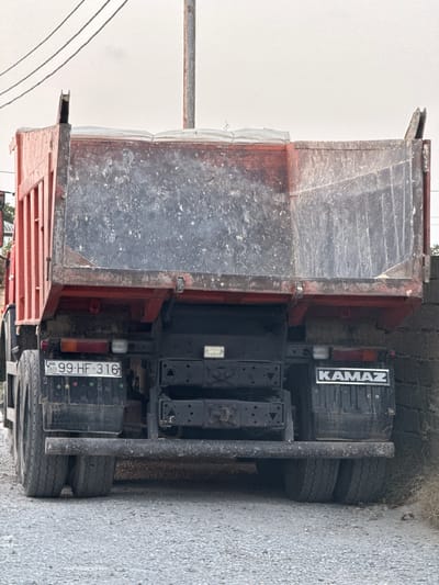 In Mastaga, Azerbaijan, a rear view of a red and black Kamaz dump truck is captured. The truck is parked on a gravel surface with a rough stone wall to its right. The truck bed, heavily used and dirty, is raised slightly, revealing the mechanisms underneath. A license plate reading "99-HF-316" is visible on the left side of the truck's rear, along with a "KAMAZ" logo on the right. Overhead, power lines stretch across a cloudy sky. The overall scene depicts a utilitarian vehicle in an industrial or rural setting.