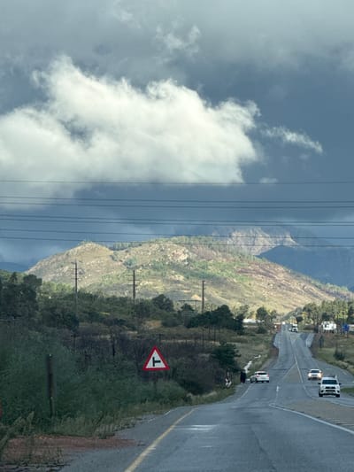 The image shows a road in Drakenstein Nu, South Africa on a cloudy day. The road is wet and has a yellow dividing line. Cars are driving on the road. A "T-junction" traffic sign is visible on the side of the road. Mountains are in the background, and power lines run across the scene. The sky is filled with dark clouds and patches of white clouds.