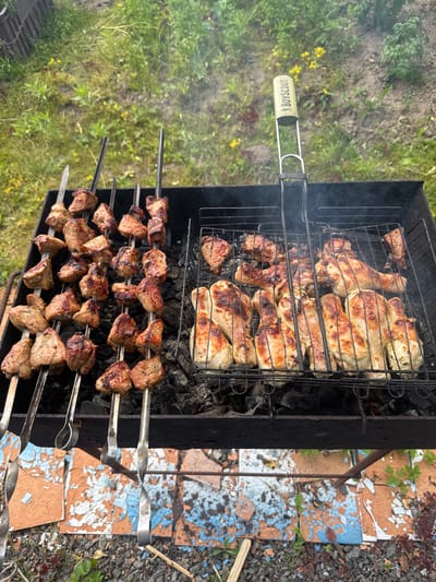 This media file captures an outdoor barbecue scene in Mazalava, Belarus. The primary subjects are various cuts of meat being grilled over hot coals in a black metal barbecue grill. On the top rack, chicken pieces, including drumsticks and possibly other parts, are arranged, some appearing well-browned and crispy. Below them, on multiple skewers, are pieces of marinated meat, likely pork or lamb, threaded tightly and showing signs of being grilled to a juicy finish. The skewers are arranged in rows, with several tiers of meat evident.
The setting is an outdoor area with a backdrop of green grass and some scattered yellow wildflowers, suggesting a rural or park-like environment. The ground around the grill is covered with small stones and some broken, colorful tiles, hinting at a rustic or perhaps a slightly dilapidated outdoor cooking area. Wisps of smoke rise from the grill, indicating that the cooking is in progress. The lighting suggests it is daytime, with natural light illuminating the scene.
There is visible text on the handle of a barbecue tool on the left side of the image. It reads "BOY SCOUT" in yellow lettering on a white background.
The overall impression is one of a casual outdoor meal preparation, likely a gathering or a recreational activity, with the focus on the process of grilling appetizing food. The image evokes a sense of warmth and the anticipation of a delicious meal.