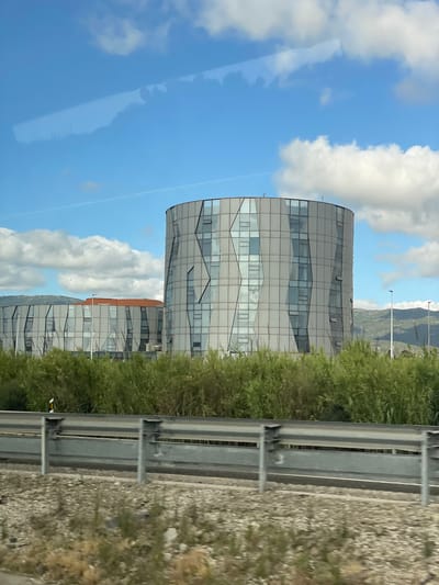 This media file captures a view of a modern, curved building with a glass facade. The building appears to be multi-story, with distinct geometric patterns formed by the window panes and structural elements. A second, similar building is partially visible behind the first. The scene is dominated by a bright blue sky with scattered white clouds, suggesting a clear and sunny day. In the foreground, there is lush green vegetation and a metal guardrail, hinting at a roadside or highway setting. The background reveals rolling hills or mountains, characteristic of a scenic landscape. The time of day appears to be daytime, likely late morning or afternoon, given the brightness of the sun. There is no visible text in the image. The overall impression is one of contemporary architecture integrated into a natural environment. The context provided places this scene in Los Barrios, Spain.