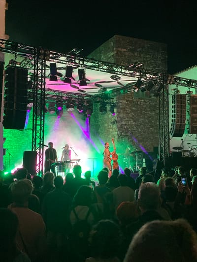 The image depicts a nighttime outdoor concert scene in Loulé, Portugal, likely within the historic old town due to the stone walls visible in the background. A band is performing on a stage illuminated by vibrant green and purple lights. On stage, three musicians are visible: a man standing behind a keyboard, a woman playing a double bass, and another person, possibly a drummer, seated behind a drum kit. The stage is supported by a metal truss structure, adorned with numerous spotlights and large speaker arrays. In front of the stage, a crowd of spectators is gathered, silhouetted against the stage lights, with some individuals holding up phones, presumably to record the performance. The overall atmosphere is energetic and festive, typical of an outdoor music event.