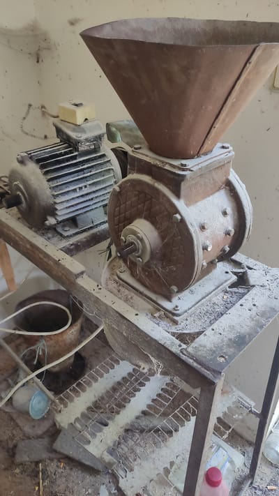 The image shows a dusty, disused industrial machine, possibly a grinder or shredder, in what appears to be a storage or workshop area. The machine is made of metal and has a large conical hopper at the top, leading to a motor. There are also visible metal grates and pipes. The overall scene is cluttered and suggests a lack of recent use, with dust and cobwebs present on the equipment. In the background, a plastic bottle with a red cap and another bottle with a white cap are visible. The setting appears to be indoors, given the lighting and the presence of what looks like a light fixture on the ceiling. There is no discernible activity or event taking place; the machine is stationary. No people are visible in the image. The location context is Mohyliv-Podilskyi, Ukraine. There is no visible text that can be deciphered.