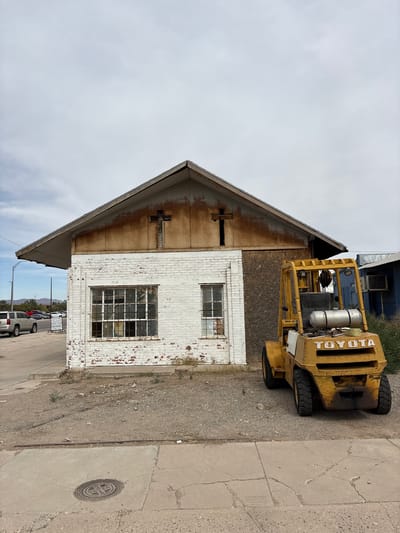 In Lordsburg, United States of America, a weathered, white-brick building with a rusted metal roof and two cross-shaped openings stands under an overcast sky. To its right, a yellow Toyota forklift is parked on a gravel lot. The building features a large window with a grid of panes on the left and a smaller window to its right. In the background, a blue building and distant mountains are visible. The foreground shows a concrete sidewalk and a circular metal grate embedded in it.