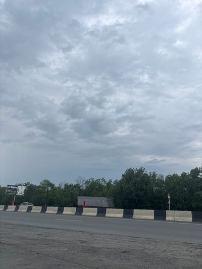 The image depicts a cloudy sky above a highway in the city of Gizel, Russia. On the left side of the highway, a billboard with Russian text and an image of food is visible. A car is parked behind it. A concrete barrier with alternating black and white sections separates the highway from the trees lining the road. A truck is stopped behind the barrier, and a triangular road sign indicating a hazard is also present. The road surface is asphalt with gravel.