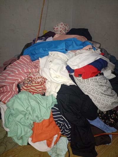 The image shows a large pile of laundry in a dimly lit room, likely a domestic setting in Blangcotbaroh, Indonesia. The laundry is a mix of colors and patterns, including striped shirts, a blue garment, a red shirt, and a white shirt. There are also some patterned items, such as a fabric with small white circular designs and another with a floral print. A thin, coiled wire, possibly an electrical cord, is visible running vertically through the pile of clothes. The floor beneath the laundry appears to be made of wood planks, suggesting an indoor environment. The lighting is poor, creating shadows and making it difficult to discern specific details about the clothes or the surroundings. There is no visible text in the image. The scene depicts a common household chore or a state of disarray, with no indication of any particular event or time of day other than the low light suggesting it might be evening or the room lacks adequate illumination. The overall impression is one of everyday life and the mundane task of dealing with laundry.