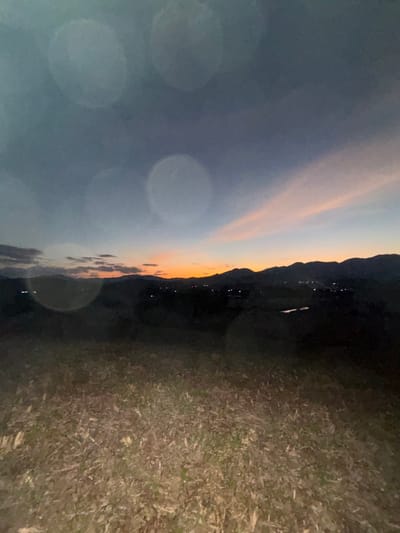 The image captures a panoramic landscape at dusk, observed from an elevated vantage point overlooking Ban Tung Yao, Thailand. The sky, occupying the upper portion, transitions from a deep blue-grey overhead to a horizontal band of orange and pink along the western horizon, characteristic of sunset. Several prominent, semi-transparent circular lens flares are present in the upper and mid-sections of the sky. A chain of dark, silhouetted mountains defines the distant background. In the mid-ground, below the mountain line, scattered clusters of small, illuminated points of light indicate the settlement of Ban Tung Yao. A dark, reflective body of water is discernible within this mid-ground, slightly right of center. The foreground consists of a vast expanse of low-lying, dry, light-brown vegetation interspersed with darker patches of earth, occupying approximately the lower two-thirds of the frame. No human subjects, distinct structures other than the distant lights, or specific actions are identifiable.