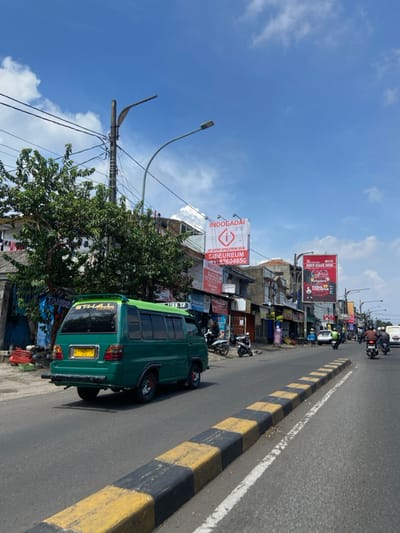 The image shows a street scene in Kecamatan Cimahi Selatan, Indonesia, at the coordinates -6.899 latitude and 107.562 longitude. A green van with the word "THALL" in white lettering on the side is driving down the road. There are parked motorcycles and a few pedestrians on the sidewalk. There is a large billboard on the left side of the street with the words "INDOGADAI" and "CIBUREUM" on it. There is another billboard on the right side of the street with a red image of a person playing a musical instrument. The street is lined with buildings on both sides, with shops and businesses on the ground floor. The sky is blue with a few white clouds. The pavement is black with yellow stripes.