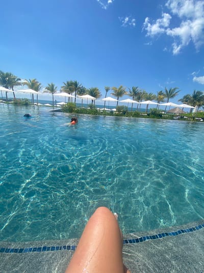 The image captures a scene at a pool in Marina Maroma, Mexico. A person's leg is visible in the foreground, resting against the pool's edge, which is lined with blue mosaic tiles. The pool water is clear and reflects sunlight. In the background, a row of white sun umbrellas lines the pool, with palm trees beyond them. Two people are swimming in the pool. The sky is blue with scattered white clouds.