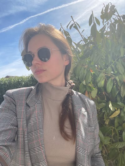 A young woman wearing sunglasses and a plaid blazer stands outside on a sunny day. She is positioned in front of a green leafy bush with the blue sky and contrails visible above. It is not possible to determine the location of the city of Kranj, Slovenia.