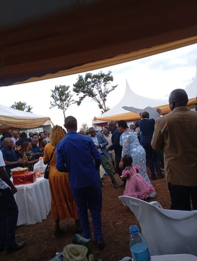 This image captures a lively outdoor celebration, likely in Shimbi Mashariki, Tanzania, with numerous people gathered under a large canopy. The scene suggests a festive event, possibly a wedding reception, birthday, or engagement, given the presence of gifts, celebratory attire, and dancing.
In the foreground, a dark awning or canopy partially covers the top of the frame, providing shade. The ground is a mix of dirt and sparse grass. Many adults are visible, dressed in various outfits, including a man in a blue suit, a woman in a patterned light-colored dress, and another woman in a mustard yellow dress. Several individuals appear to be clapping, dancing, or interacting, creating a dynamic and joyful atmosphere. A small child, wearing a ruffled pink dress, is a prominent figure in the mid-ground, seemingly dancing or moving through the crowd.
To the left, a table covered with a white tablecloth holds several wrapped gifts in red and gold. In the immediate foreground, part of a white chair and a plastic water bottle are visible, along with what appears to be white artificial roses on a surface.
In the background, large white peaked tents, adorned with gold or orange fabric draping, define the outdoor venue. Two prominent trees with green foliage stand against an overcast, light sky, indicating either late afternoon or an overcast day. No discernible text is visible in the image. The overall impression is one of communal celebration and happiness.
