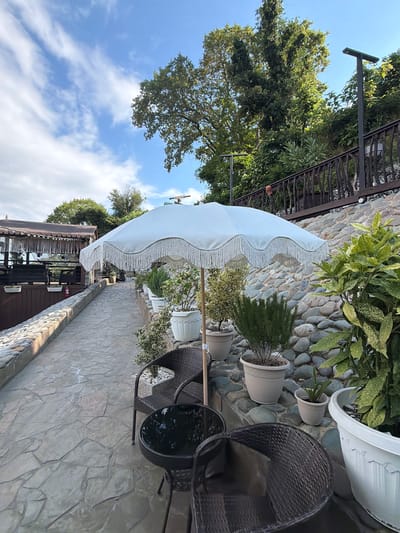 The image depicts an outdoor seating area at what appears to be a restaurant or cafe in Nizhnyaya Shilovka, Russia. A white fringed umbrella shades a stone-paved walkway lined with potted plants. Two wicker chairs and a small dark table are positioned near the walkway. In the background, a building with a wooden deck and thatched roof is visible. The area is situated on a hillside covered with lush green trees and a stone wall, suggesting a scenic location. The sky is blue with scattered white clouds. No people are present in the image.