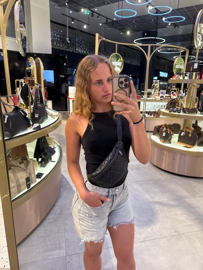 A young woman with blonde, curly hair is taking a mirror selfie in a brightly lit luxury handbag store. She is wearing a black halter top and light-wash denim shorts with frayed hems. A black quilted fanny pack is slung across her chest. The store interior features polished floors, illuminated display shelves showcasing various handbags, and decorative gold accents. In the background, several round mirrors with gold frames are visible, reflecting the store's lighting and merchandise. One of the mirrors prominently displays the "DKNY" logo. The woman is holding a smartphone in her right hand, angled to capture her reflection. The scene appears to be inside a retail establishment within the city of Surcin, Urban Municipality, Serbia, likely a shopping mall given the modern interior design and wide selection of designer goods.