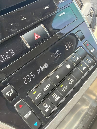 The image presents a close-up view of a car's central console, focusing on the climate control and media interface, set in Umm Salal Ali, Qatar.

The digital display prominently shows an outside temperature of 43°C, indicating extremely hot weather, consistent with the region. The cabin's set temperature is displayed as 23.5°C, suggesting the air conditioning system is actively cooling the interior. A fan speed indicator shows a medium-high setting, and the 'AUTO' climate control function is illuminated green. The time displayed is 0:23.

Key controls visible include 'TEMP' buttons with red and blue arrows for temperature adjustment, buttons for 'A/C', 'DUAL' climate zones, 'AUTO', and 'OFF'. Airflow distribution buttons are also present, including those for 'FRONT' and 'REAR' defoggers, along with icons for various cabin vents and recirculation.

Below the climate controls, there are 'VOL' buttons with '+' and '-' for audio volume adjustment, an 'EJECT' button, and text indicating 'USB/SBB' input options. A red triangular hazard warning button is centrally located. The text 'PASSENGER' is also visible on a screen area, likely related to passenger-side climate or information display. The console surface is black plastic, showing some dust and reflections from bright ambient light.