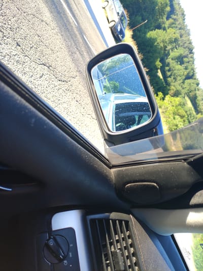 The image shows a view from inside a vehicle, looking out the side window and in the side-view mirror. The car appears to be on a road next to a line of trees. In the side-view mirror, another car is visible driving in the same direction. The interior of the car is also visible, including a dashboard with a dial and an air vent. The location is identified as Taran, Bulgaria, though the image itself does not contain any specific visual cues to confirm this location.