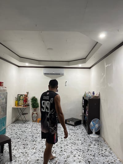 In a room with textured, off-white walls and a patterned tiled floor, a man wearing a black tank top with the name "DASCIL" and the number "99" is standing and facing away from the camera. An air conditioning unit is mounted high on the wall above him. To the left, a small table holds a framed picture of Jesus, a small statue of a child, and a potted artificial plant. Further left is a multi-tiered cabinet. To the right, a dark wood shelf unit displays various bottles and containers. In front of the shelf unit, a white oscillating fan is running. A black electronic device, possibly audio equipment, is on the floor near the center of the room. The ceiling is white with dark trim. The overall impression is of an indoor domestic setting.