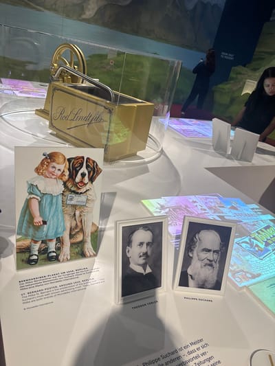 The image shows a museum display, likely in Kilchberg (ZH), Switzerland, given the presence of chocolate brands produced there. The display features a glass case containing a gold-colored "Rod Lindt fils" carriage. Next to the case are framed black and white portraits of Philippe Suchard and Theodor Tobler. A poster of a young girl with a St. Bernard dog advertises "Milka" chocolate. The display showcases the history of the Milka chocolate brand and its connection to Suchard and Tobler. In the background, a screen shows an image of two people with text that reads, "FROM PAST TO PRESENT."