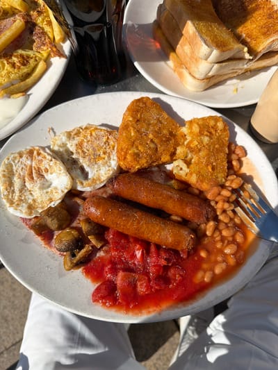 The image displays an outdoor meal setting, likely a breakfast or brunch, under strong sunlight, indicated by sharp shadows. The scene is observed from a high angle, looking down onto a table.
In the foreground, a light-colored, possibly ceramic, plate contains a substantial meal. Components include:
- Two fried eggs, sunny side up, with fully set whites and intact yolks.
- Two rectangular hash browns, one of which is partially broken, revealing its shredded potato interior.
- Two cooked sausages, browned and plump.
- A portion of baked beans in a red-orange tomato sauce.
- A separate serving of chopped or stewed red tomatoes.
- Several sautéed button mushrooms.
A silver-colored fork with four tines is partially visible on the right side of this plate, resting within the baked beans.
In the background, three additional items are present:
- To the left, a white plate holds a yellow, possibly eggy or pancake-like dish, accompanied by what appears to be fried potato strips or similar elements, and a dollop of white sauce.
- Between the left background plate and the main foreground plate, a dark glass bottle, consistent with a cola bottle, stands upright, showing condensation, indicating it is a cold beverage.
- To the right, another white plate is stacked with several slices of lightly toasted white bread.
- A beige or cream-colored, cylindrical object, possibly a condiment container, is partially visible on the far right edge of the frame.
The lower portion of a person's legs, clad in white trousers, is visible beneath the main foreground plate, suggesting the individual is seated at the table. The ground surface visible beneath the legs and around the table appears to be a paved or hard-surfaced area, consistent with an outdoor patio or seating area.
The specific location within Crewe, United Kingdom, is not visually identifiable from the provided image details.